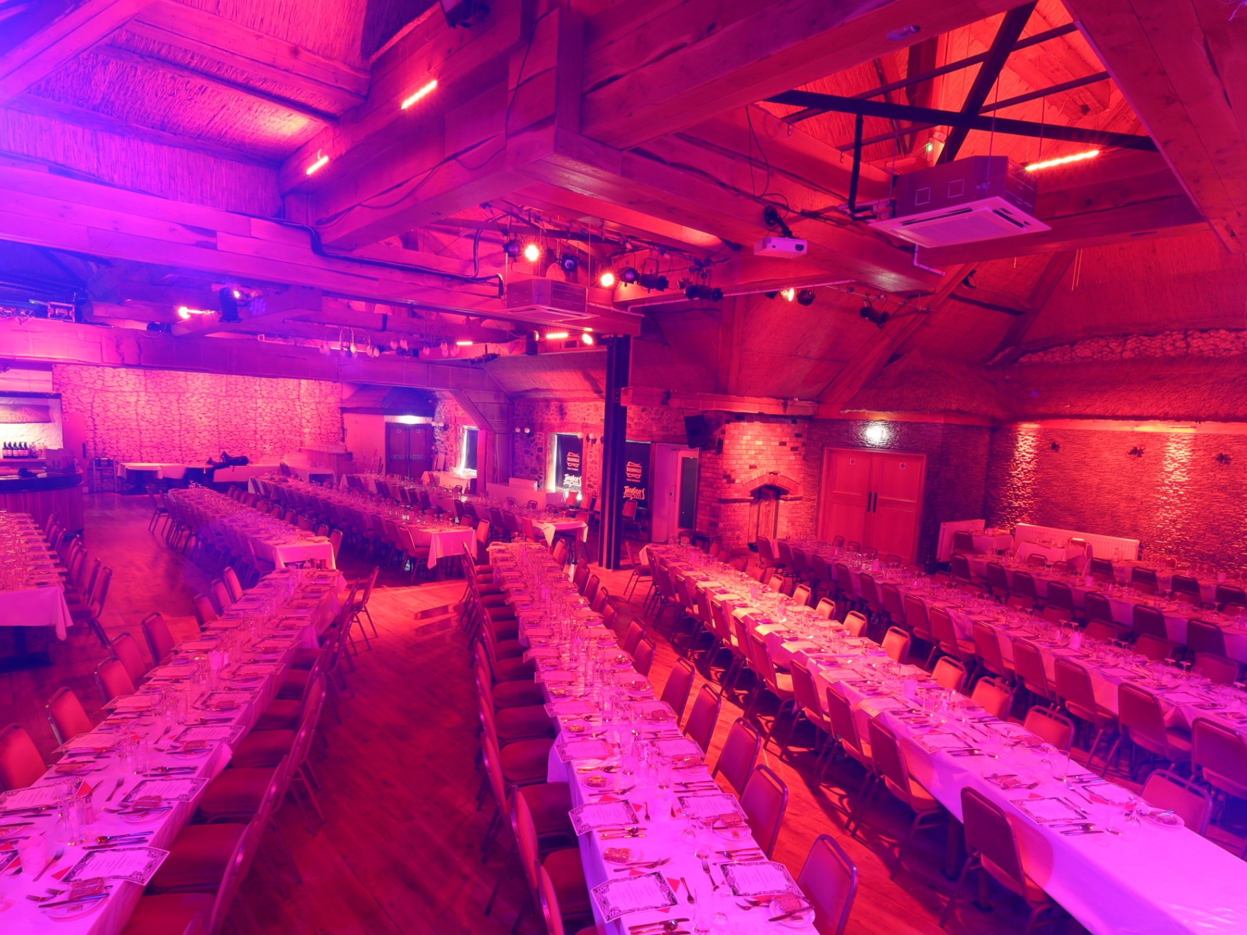 Large hall with long tables set for a banquet, lit in pink and purple tones.