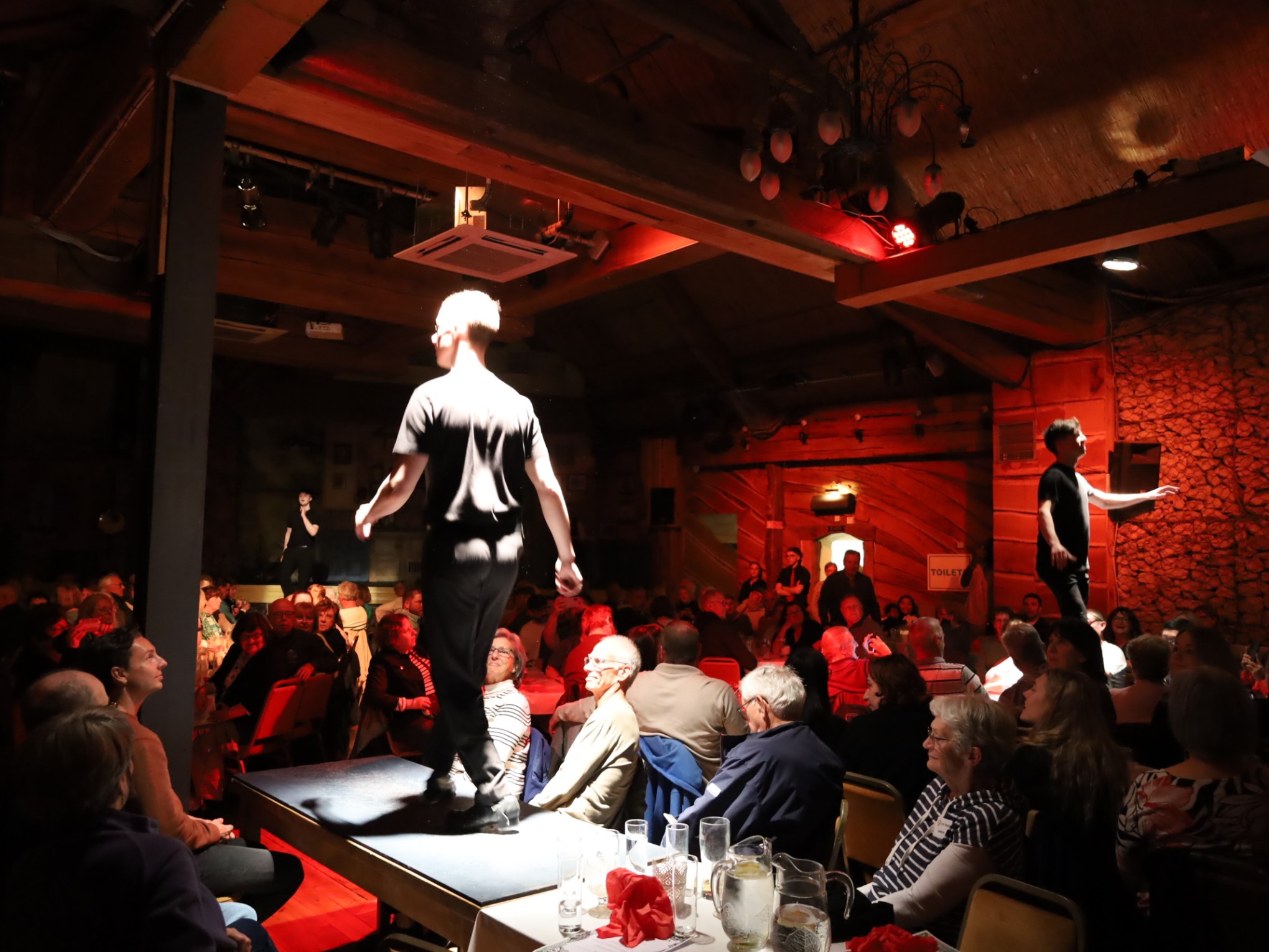Two performers on stage with an audience seated in a dimly lit venue.