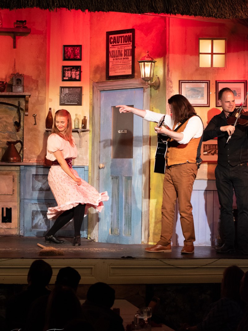 Performers in a colorful room; woman dancing, man playing guitar, another with violin.
