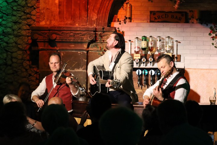 Three musicians perform on stage with guitars and a violin in a dimly lit bar.