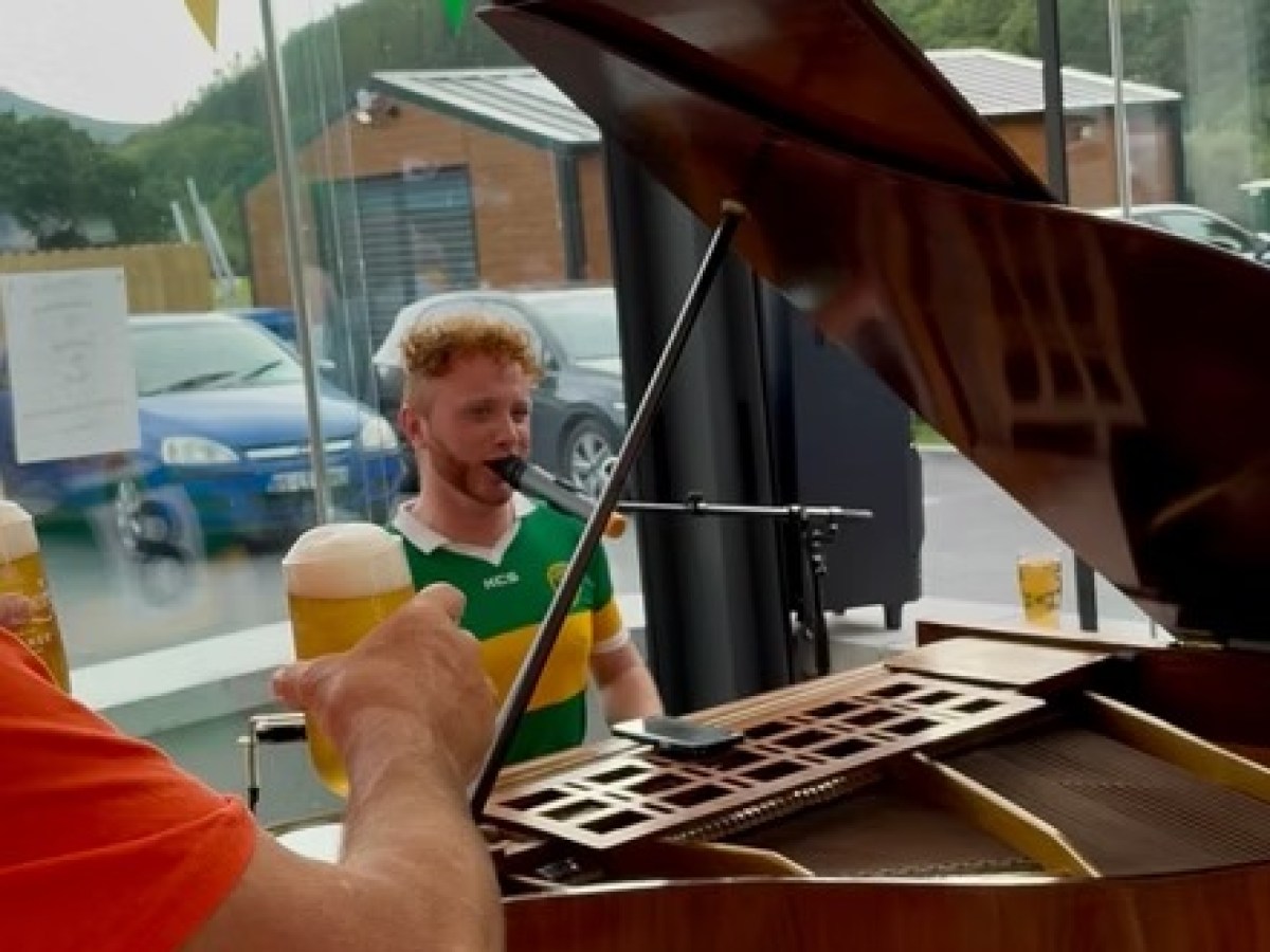 Person playing piano indoors, with glass roof, colorful banners, and a hand holding a beer nearby.