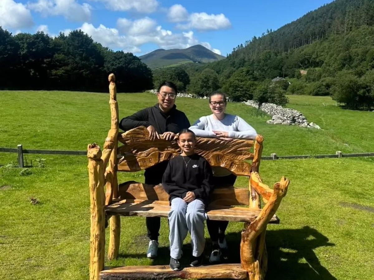 Three people sit on a large wooden chair in a grassy field with mountains and blue sky in the background.